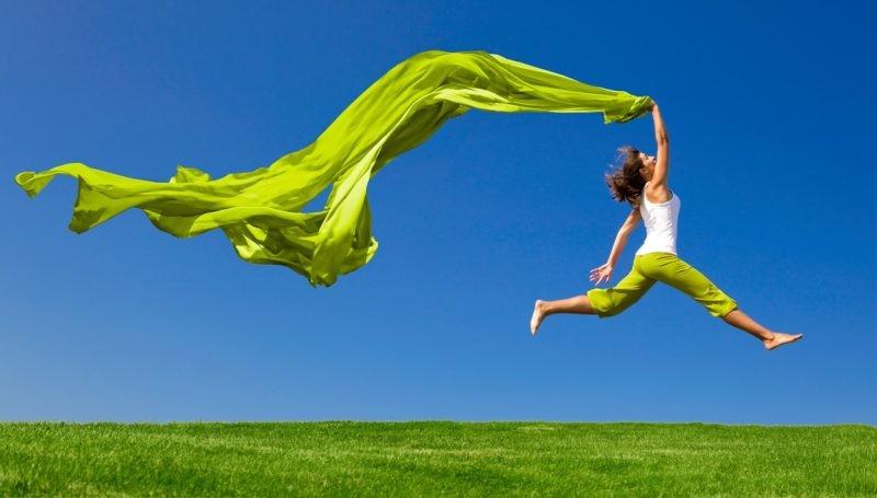 Beautiful young woman jumping on a green meadow with a colored tissue